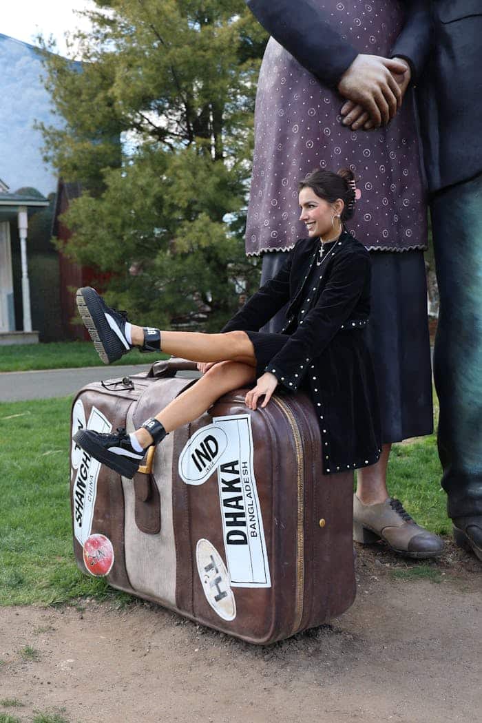 Woman seated on artistic giant suitcase in park, embracing travel theme.