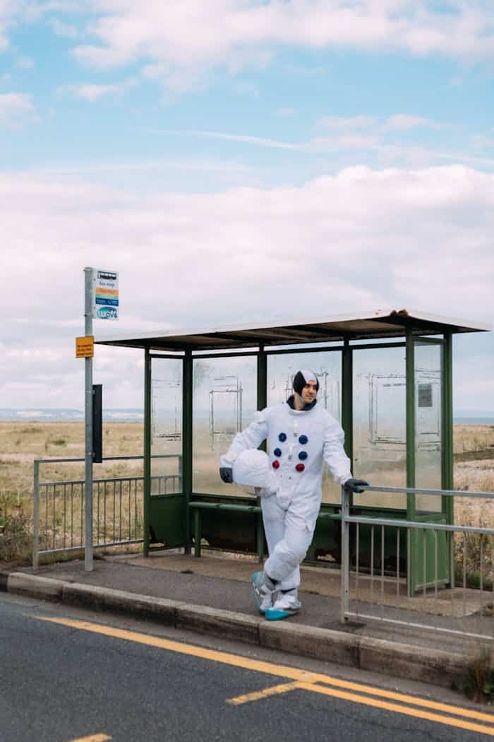 Man in a space costume standing by a rural bus stop, under a blue sky, during the day.