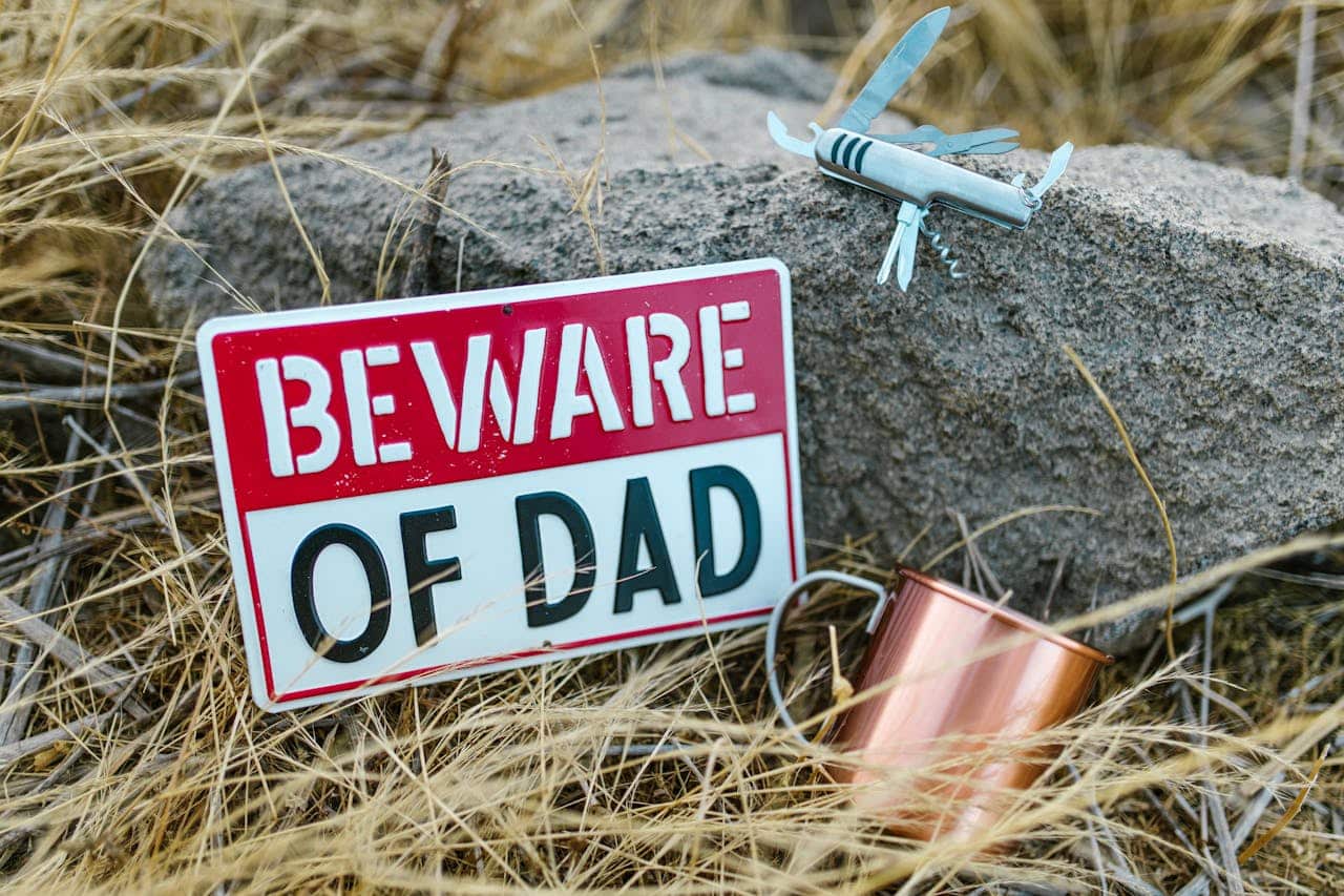 A humorous 'Beware of Dad' sign with outdoor tools in a natural setting.